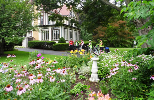 A group of bicyclists in the driveway of a Tudor mansion with flowers in the foreground.