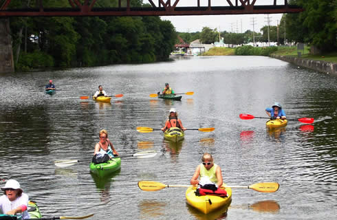 Several people are kayaking down calm water on a river under a bridge.