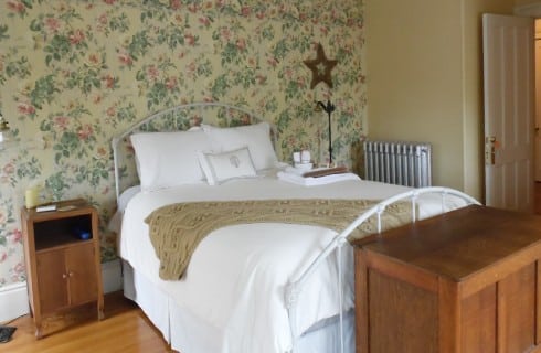 Bedroom with floral wallpaper and a bed made up in white with cherry wood furniture.