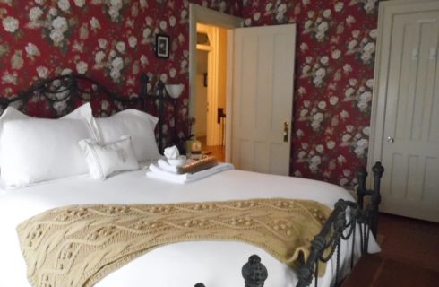 Bedroom with red floral wallpaper, white trim and a black iron bed made up with white bedding.