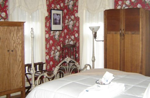 Bedroom with floral red wallpaper, wooden cabinets and a white iron bed made up in white bedding.