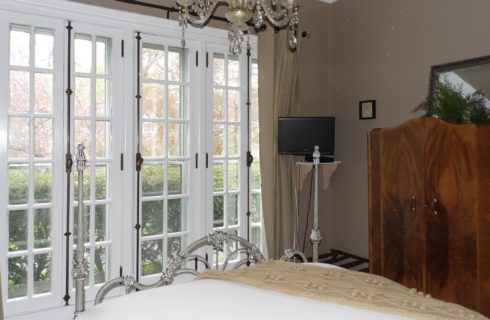 Elegant bedroom with huge french windows, an ornate iron bed, a chandelier, and an antique wooden armoire.