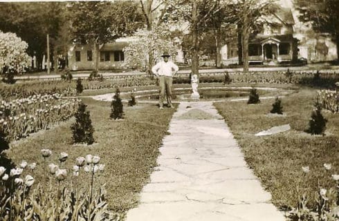 Historic sepia photo of a manstanding at the end of a arden path in front of a small pond.