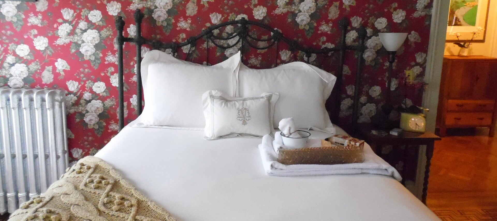 Bedroom with red floral wallpaper, white trim and a black iron bed made up with white bedding next to a white radiator.