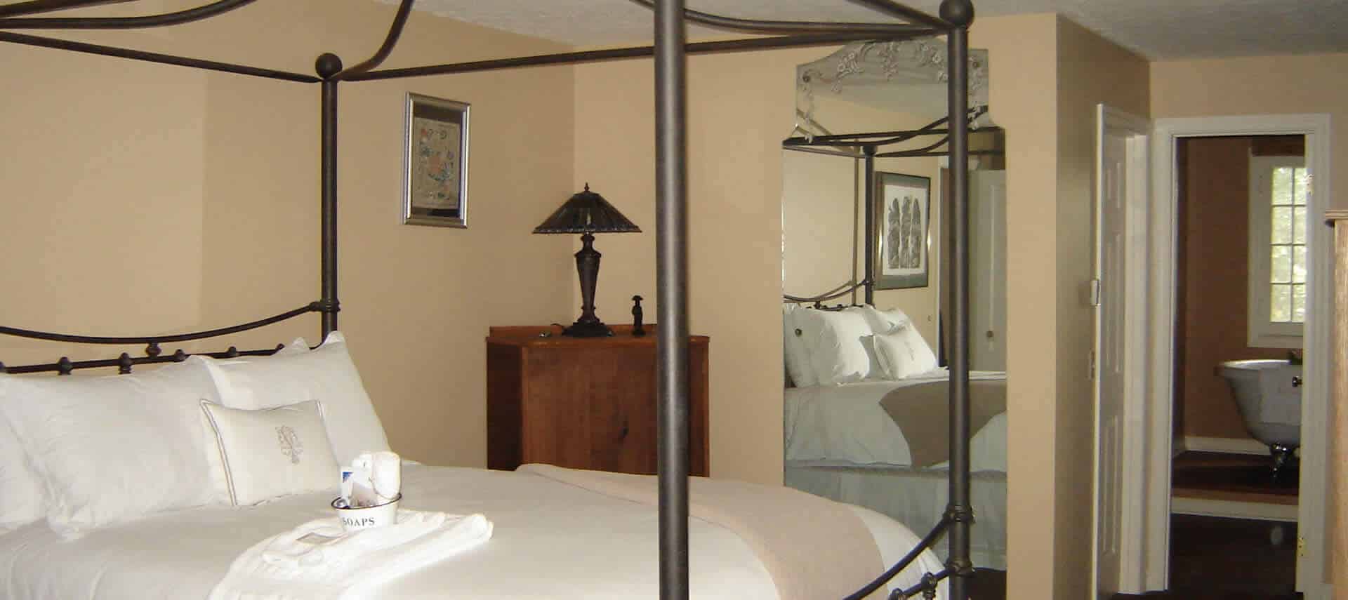 Graceful beige guestroom with a large iron canopy bed made up in white bedding, a dresser and mirror.