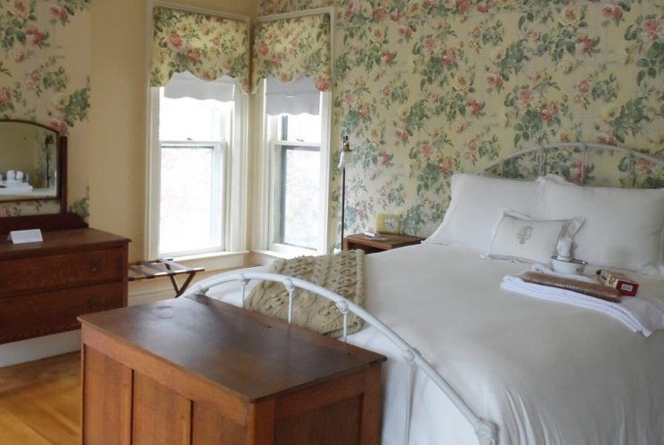 Guestroom with floral wallpaper holds an iron bed with white bedding.