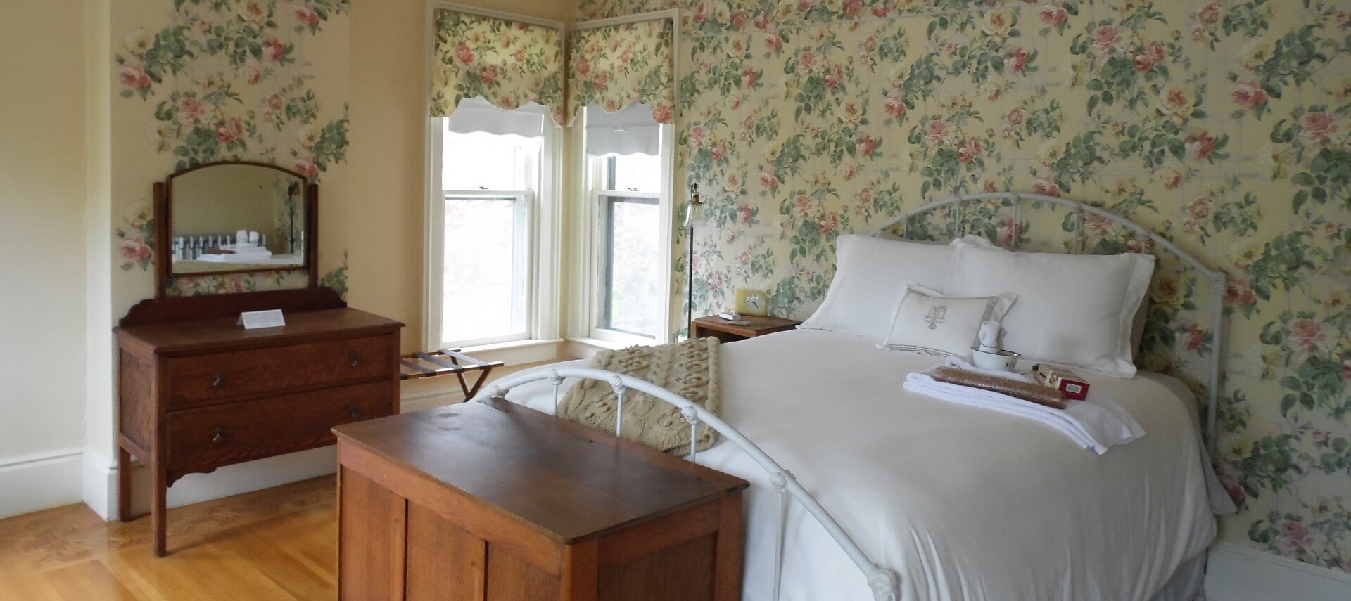 Guestroom with floral wallpaper holds an iron bed with white bedding.