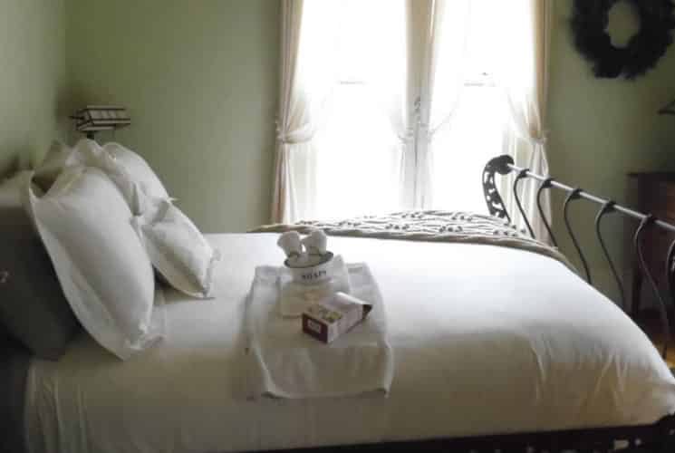 A guestroom with pale green walls holds dark wood furniture, an ornate iron bed with white bedding and a large window.