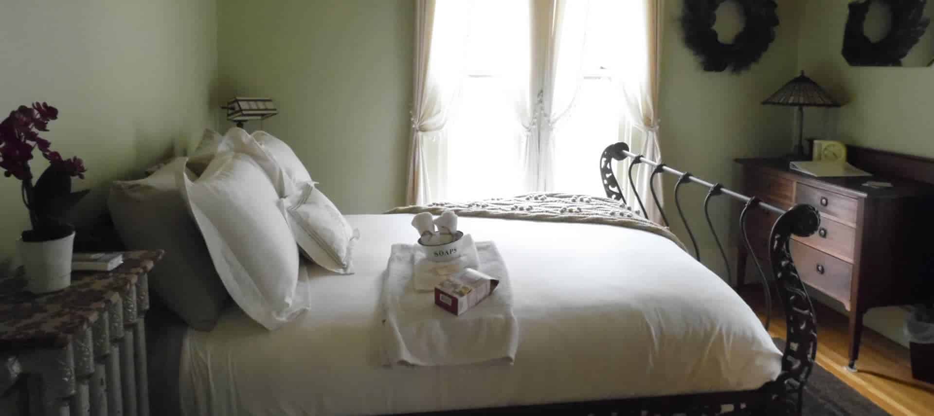 A guestroom with pale green walls holds dark wood furniture, an ornate iron bed with white bedding and a large window.