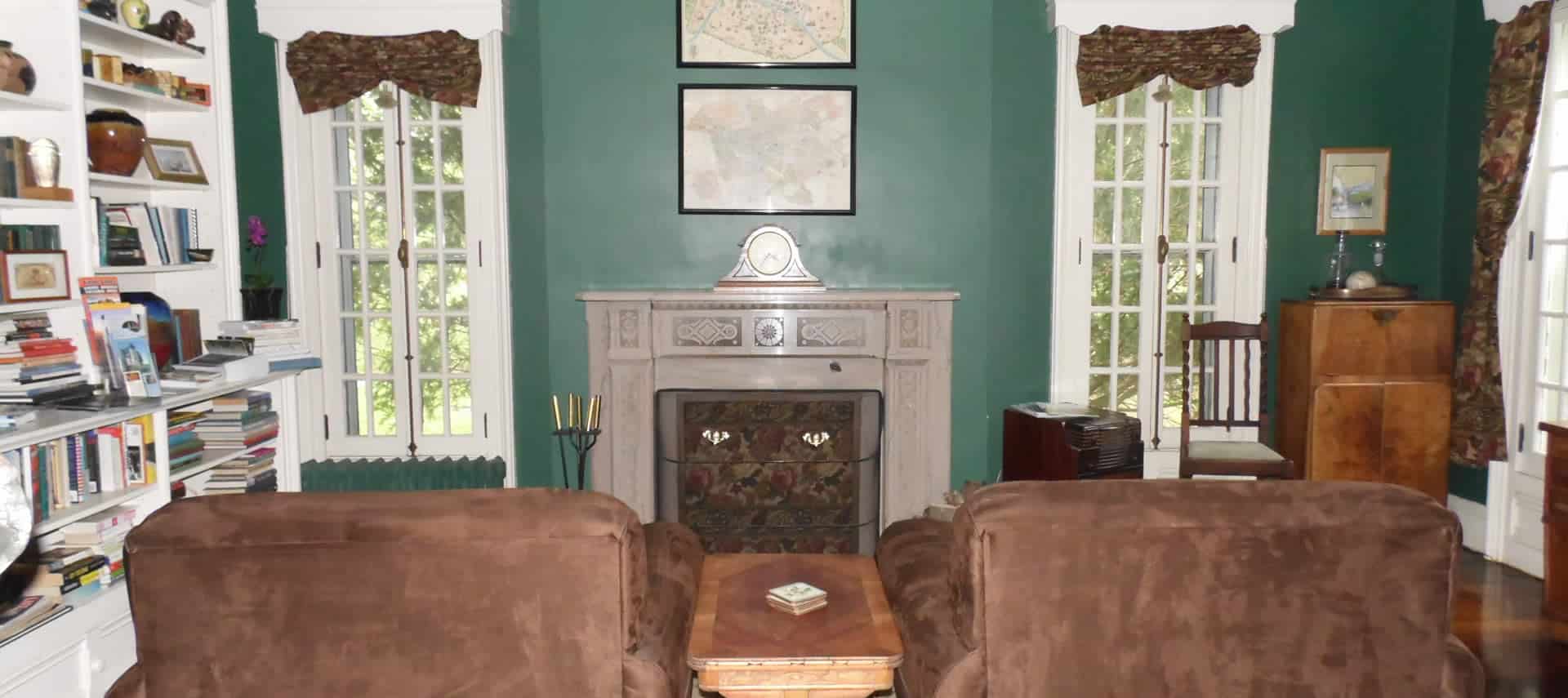 Two soft brown chairs face a white fireplace in a room with built in library shelves full of books.