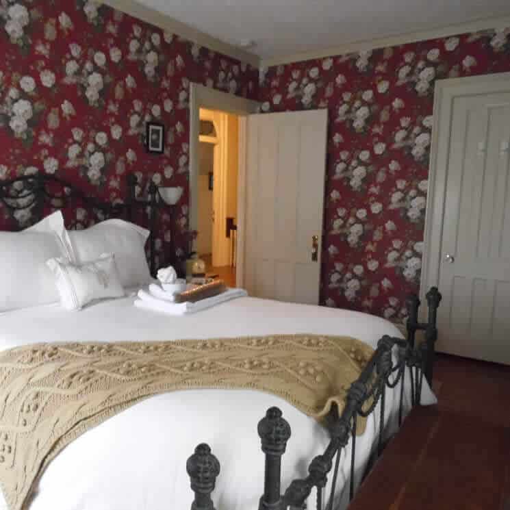 Cozy guestroom with brass bed made up in white bedding and a red floral wallpaper.