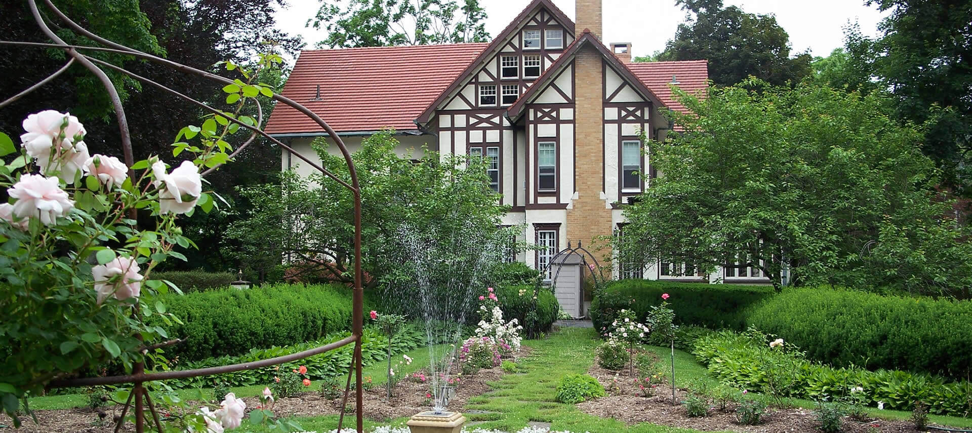 The back garden of a large Tudor mansion, with greenery and flowers. 