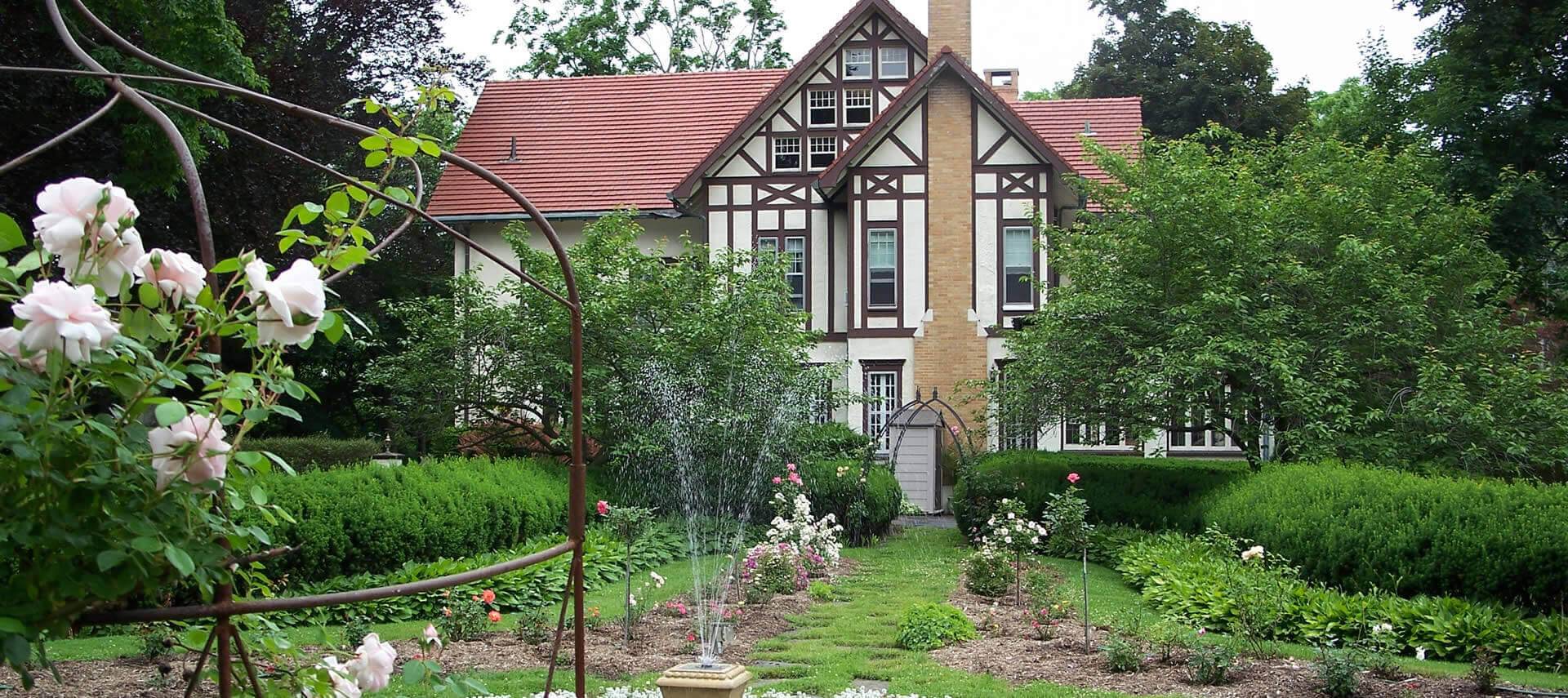 The back garden of a large Tudor mansion, with greenery and flowers. 